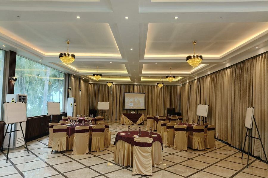 alt-text A spacious, well-lit event hall with round tables dressed in beige and burgundy tablecloths, large windows offering a view of palm trees, chandeliers, and a neatly arranged projector screen and whiteboards for a formal event.