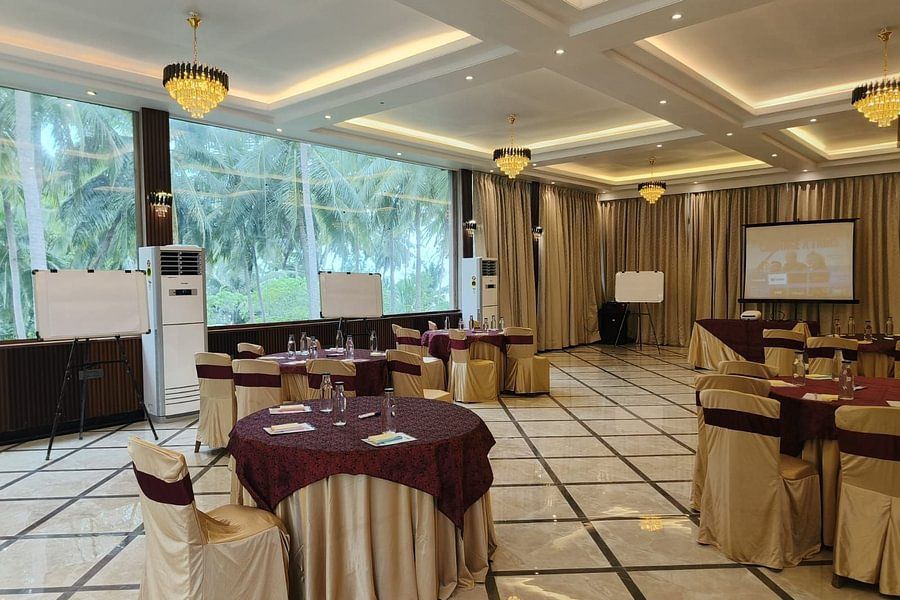 alt-text A meeting or event space featuring round tables, chairs with beige covers and burgundy sashes, chandeliers, whiteboards, and a projector screen set up in a modern hall with floor-to-ceiling windows overlooking palm trees