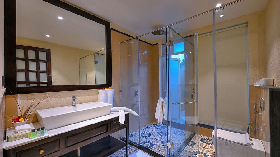 A modern bathroom of Premium Suite Cottage with a rectangular sink, a large mirror, a glass-enclosed shower with patterned tiles, a western toilet, and rolled white towels - Singhvilas, Ranthambore