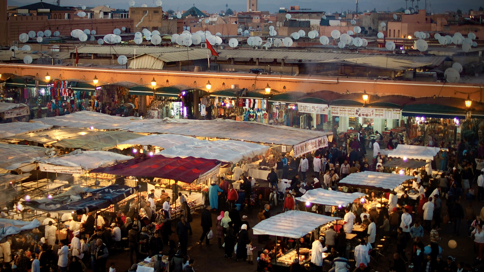 A crowded, vibrant open-air market or souk at night, filled with stalls, merchandise, and overhead lighting.