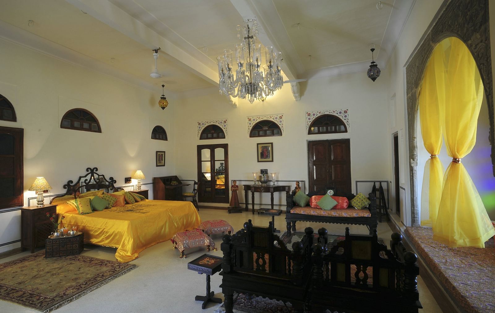 A spacious bedroom at Fort Barli, featuring a vibrant yellow bedspread, a crystal chandelier, and antique furniture.