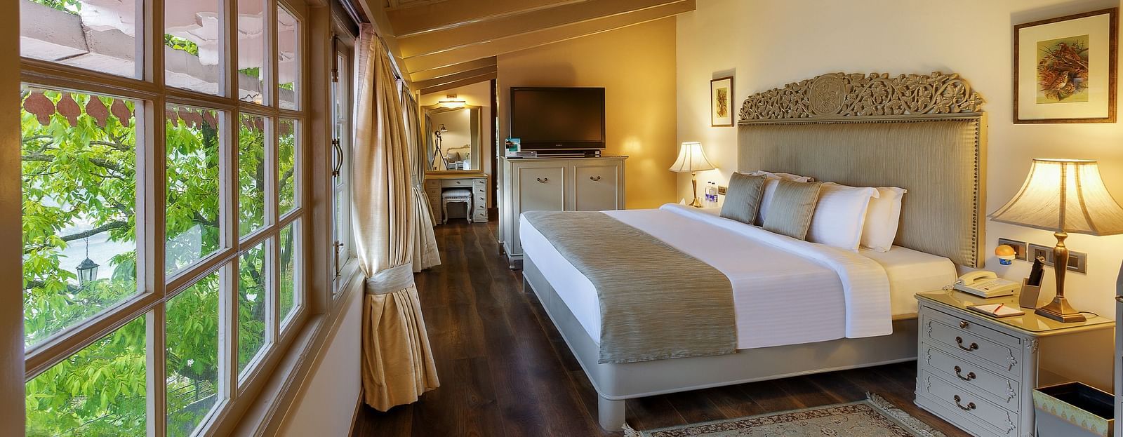 Image of a cosy room with lush decors, king size bed, wooden ceiling and  windows on the side and framed pictures on the other side- The Naini Retreat