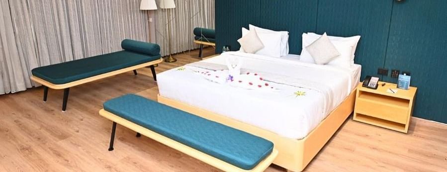 A bedroom in Dhanalakshmi Srinivasan Hotels, Perambalur, with a large bed, two lounge chairs, and two armchairs. The room features wooden flooring and a dark blue accent wall.