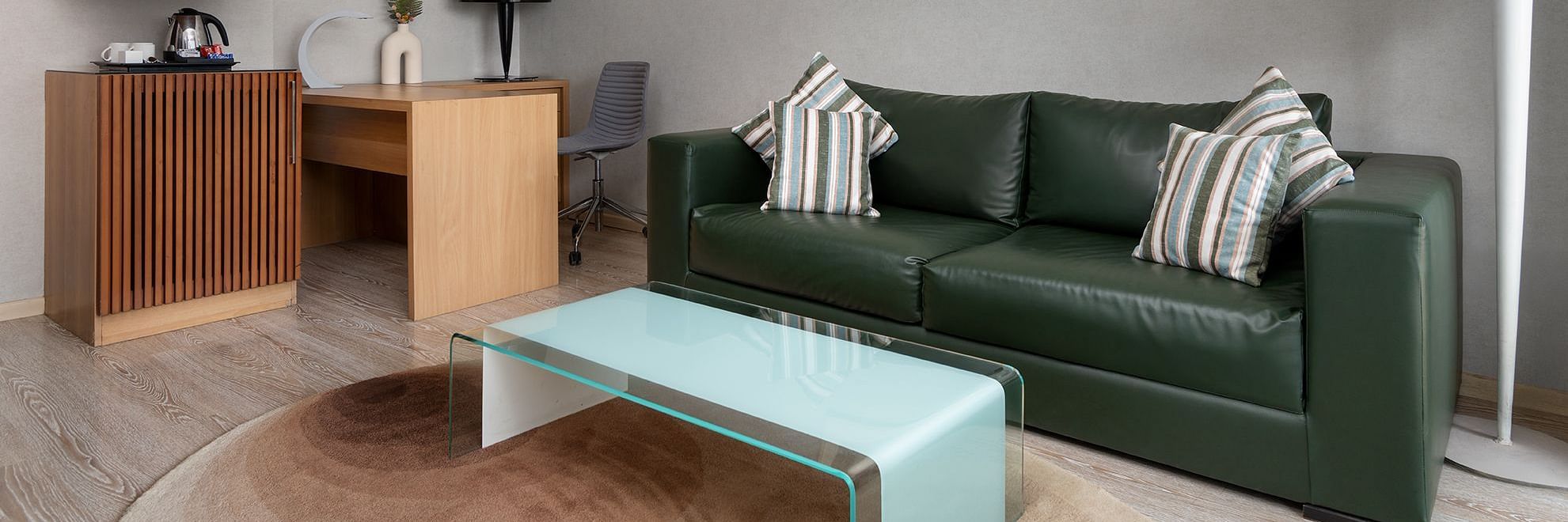 A modern room at The Atara, Gurugram featuring a green leather couch, a glass coffee table, a wooden desk with a chair, and a compact kitchenette area with a kettle