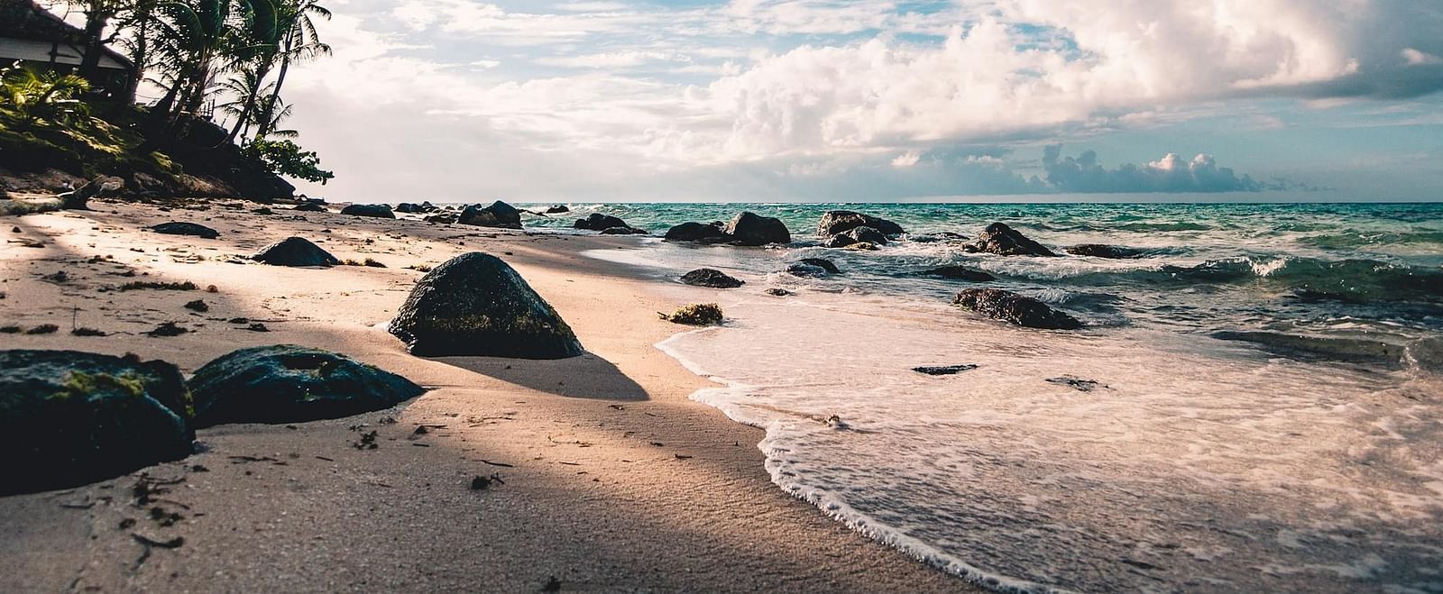A serene beach with golden sand, scattered rocks, lush palm trees, and gentle waves under a bright blue sky with white clouds.