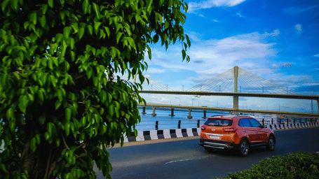 An orange SUV drives along the Mumbai coastal road beside the Bandra-Worli Sea Link under a bright blue sky framed by lush green trees.