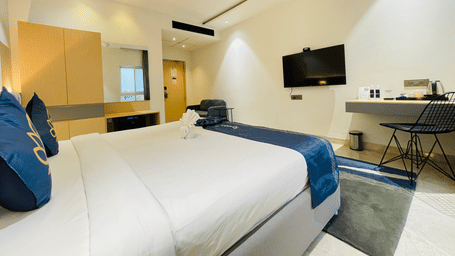 King bed room at Hotel Kyzen HiTech City with blue accent bedding, wall-mounted TV, wooden work desk, wire chair, and ceiling-mounted air conditioning unit.