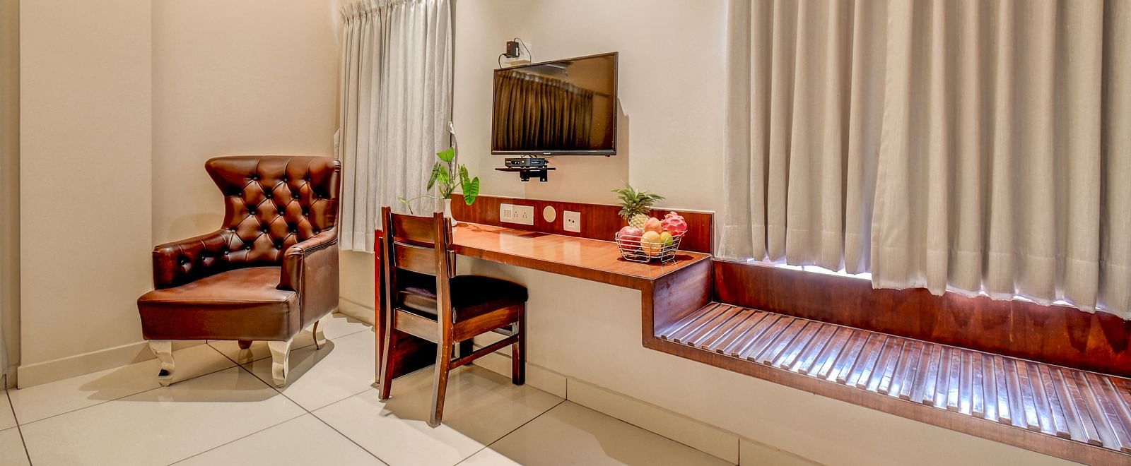 A wall-mounted TV at Harigovinda's JNStay, with a wall-mounted table below it, a cushioned chair beside, and curtains covering the window