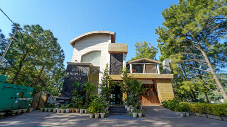 A multi-storey building with a curved roof facade and large glass windows, surrounded by tall trees, with a paved driveway leading to the entrance, lined with potted plants, and a signboard reading Hotel Whispering Winds near the doorway.