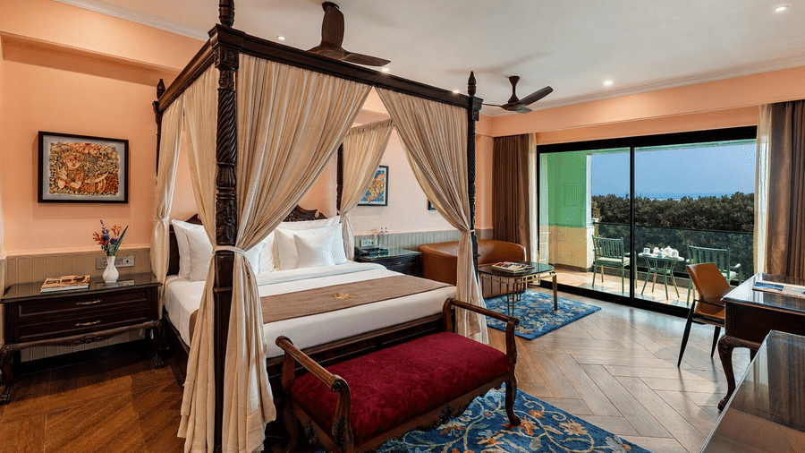 A Deluxe Suite at MAYFAIR Bay Resort, Paradeep, featuring a wooden 4-poster bed and glass doors leading to an open balcony. 