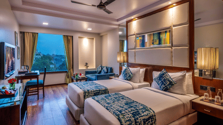 a twin bed room with a ceiling fan, wall-mounted TV, bed side table, seating area,  a workstation with a chair and half-length glass windows at Libra Lords Inn Jaipur