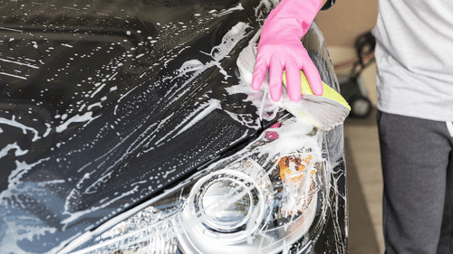 A person wearing pink gloves washes the headlight of a soapy, dark-coloured car with a yellow and white sponge