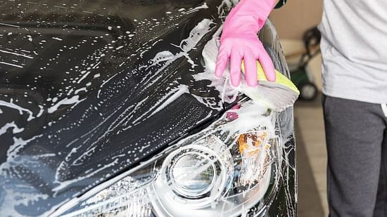 A person wearing pink gloves washes the headlight of a soapy, dark-coloured car with a yellow and white sponge