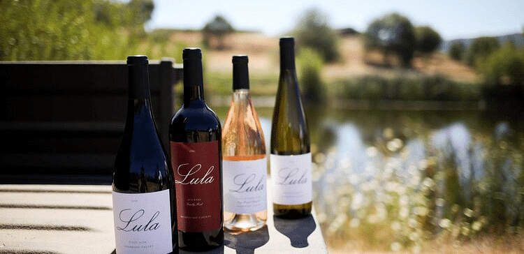 Four bottles of Lula wine stand on a table outdoors, with a blurred background of greenery and a body of water.