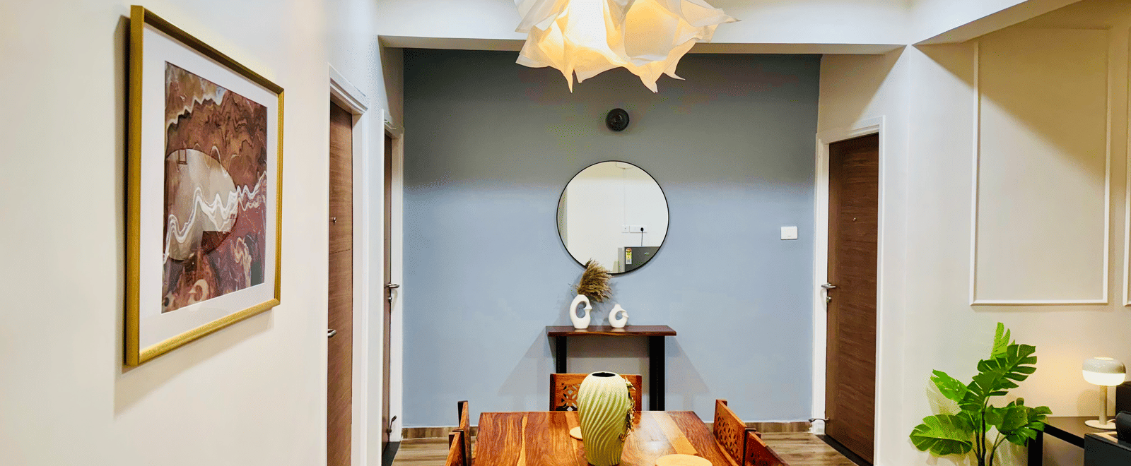 Dining area and common space in the Coral 4BHK at Kove Smart Stays featuring its wooden dining table and chairs placed at the centre of a common space that leads to doorways, and walls that are adorned with paintings and set-ups.