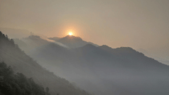 The sun rises over misty, layered mountain ridges, casting a warm golden glow through a hazy sky.