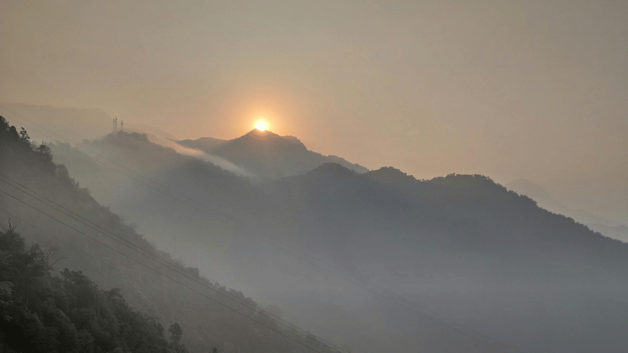The sun rises over misty, layered mountain ridges, casting a warm golden glow through a hazy sky.