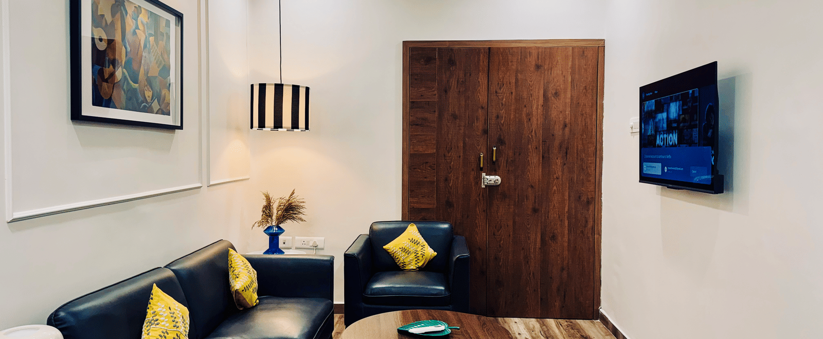 Side view of the living room of Coral 4BHK at Kove Smart Stays featuring upholstered seating areas facing a wall-mounted TV in a room with wooden flooring and ample lighting.