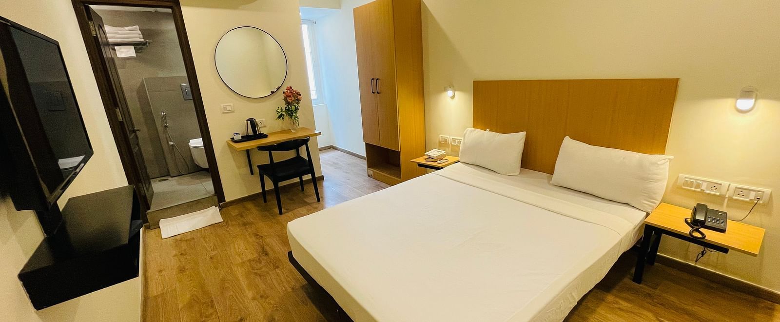 A room with a double bed, television, wardrobe, desk with a round mirror, and an open doorway - Sanctum Suites Richmond Road, Bangalore