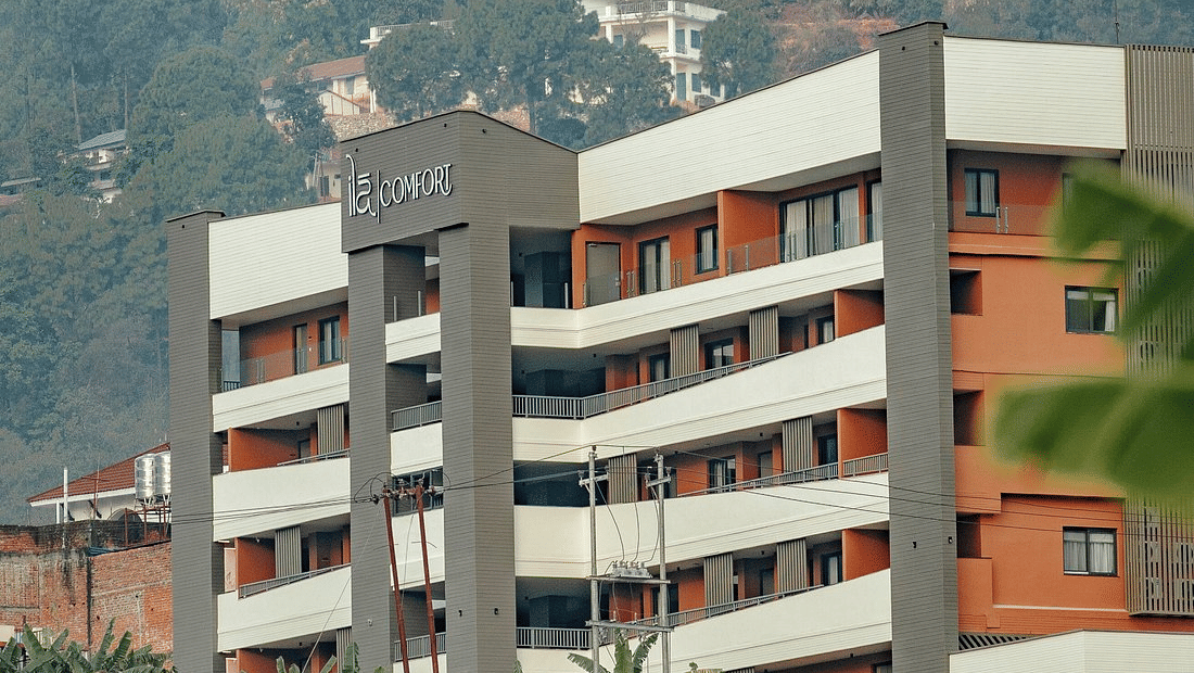 Multi-storey white and orange facade with railed balconies and rooftop brand signage set against a forested hillside at Ila Comfort, Baglung.