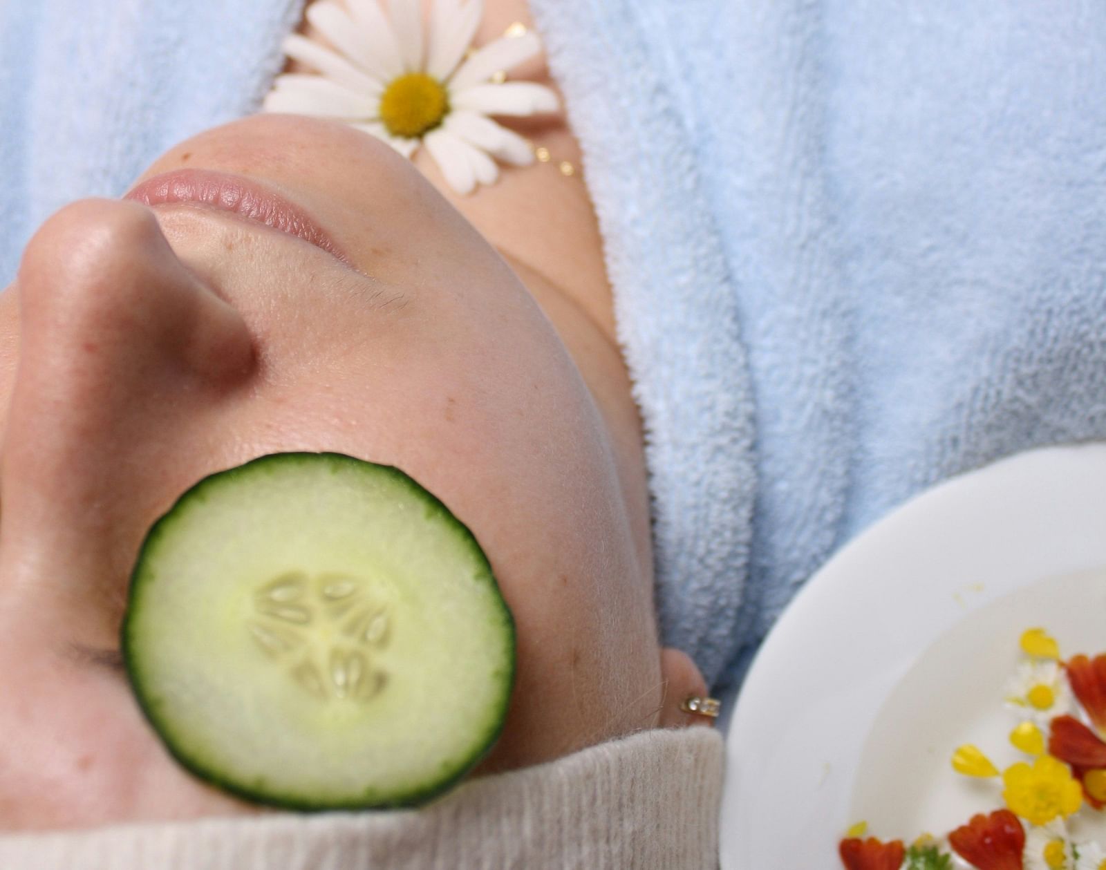 A person with a face cream and cucumber on their eye, next to a bowl of petals.