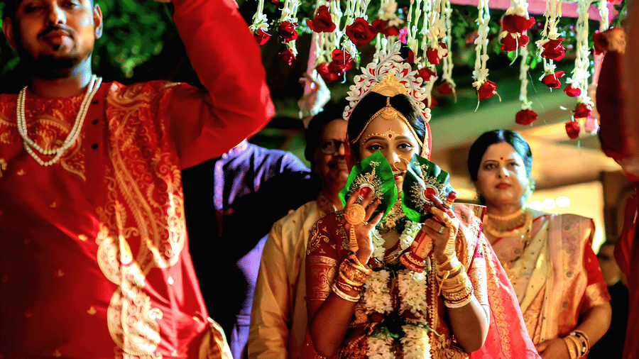 A bride and groom in traditional red wedding outfits posing together at Vedic Village Spa Resort with decorative string lights and floral garlands visible in the festive background.