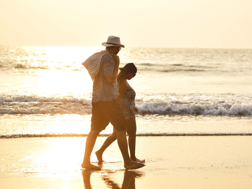 A couple walking on a wet shoreline of a beach with a bright sun reflecting the rays on the ground and the ocean.