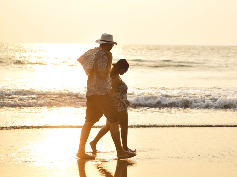 A couple walking on a wet shoreline of a beach with a bright sun reflecting the rays on the ground and the ocean.