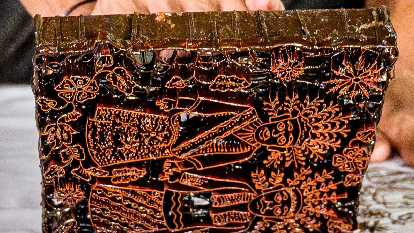 A person's hand holds a complex, rectangular wooden or copper stamp used for creating a Batik pattern, with the pattern already stamped on the white cloth below.