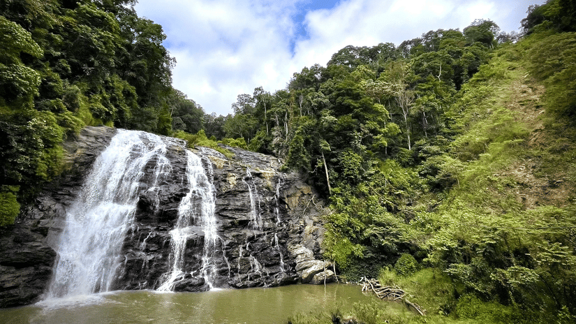 A waterfall flowing over a rocky cliff surrounded by trees and green vegetation under a partly cloudy sky.