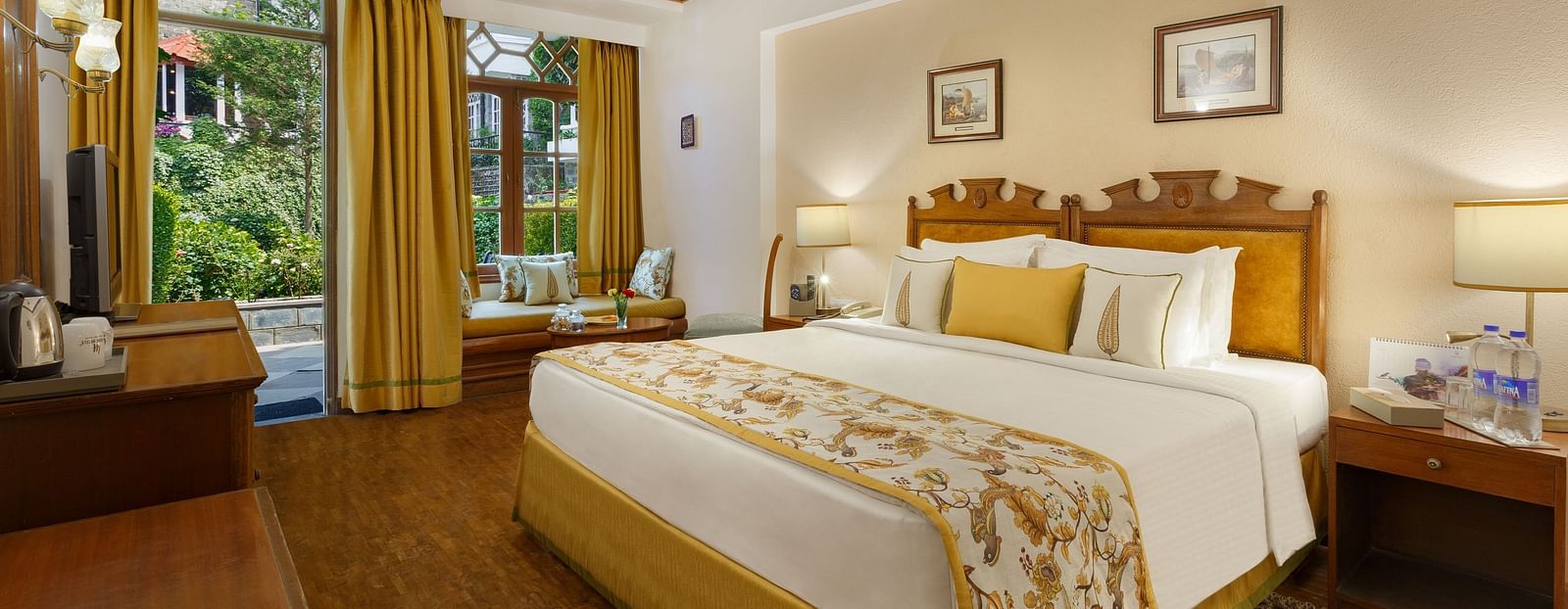 An image of a garden facing room in the naini retreat, nainital, with a king-size bed, wooden furniture, and a window. The room also features a ceiling fan, a TV and wooden flooring.