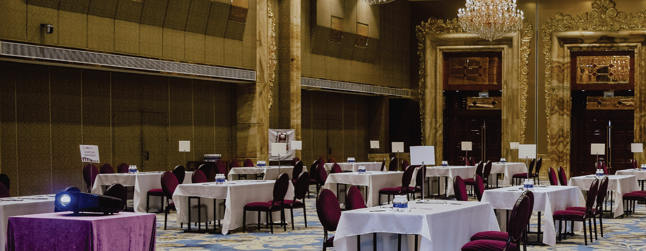 A grand ballroom features elegant chandeliers, set tables with maroon chairs, and a projector on a covered table with text - Opening Soon in Bichpuri Agra.
