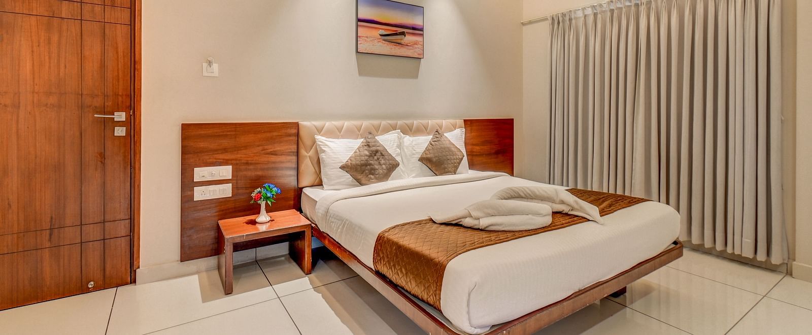 A spacious room at Harigovinda's JNStay with a large bed, wooden headboard, bedside table with a plant, a door, and curtains covering the window