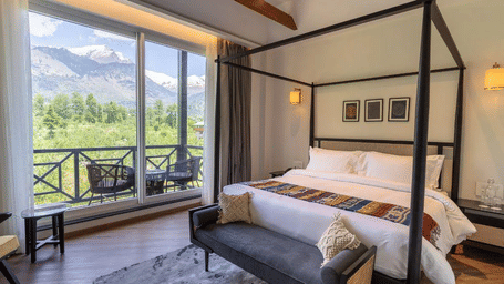 A room at Nivasana Manali featuring a four-poster bed, a couch, and a balcony with seating with stunning views of snow-capped mountains and lush greenery in the foreground.