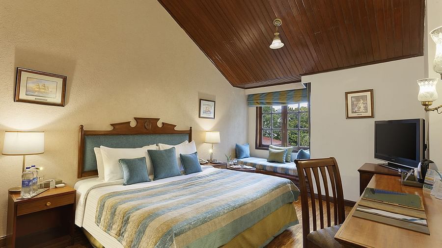 An image of a  cozy room in the naini retreat, nainital, with a king-size bed, wooden furniture, and a bay window overlooking a garden. The room features a ceiling fan, a television, and a small sitting area. 