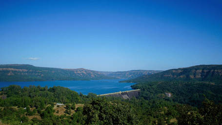 A wide landscape view of a water body surrounded by hills and dense greenery under a clear blue sky, with vegetation in the foreground.