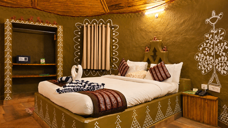 Tribal cottage bedroom with hand-painted tribal wall art, a bed dressed in patterned linen, a thatched ceiling and a curved mud wall design.