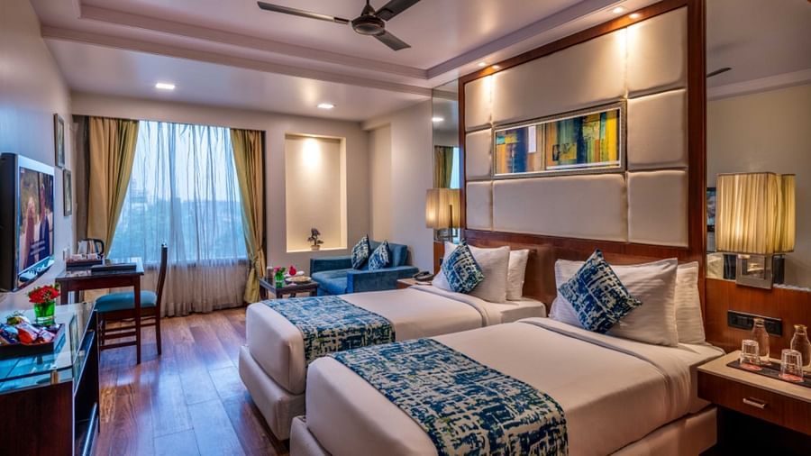 A comfortable hotel room offers two single beds, a desk with a chair by a window, a television, and a ceiling fan.