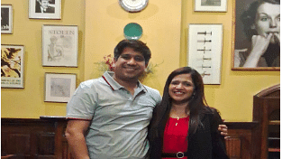 Man and woman posing together and smiling inside a warmly lit room with vintage decor and framed photos.