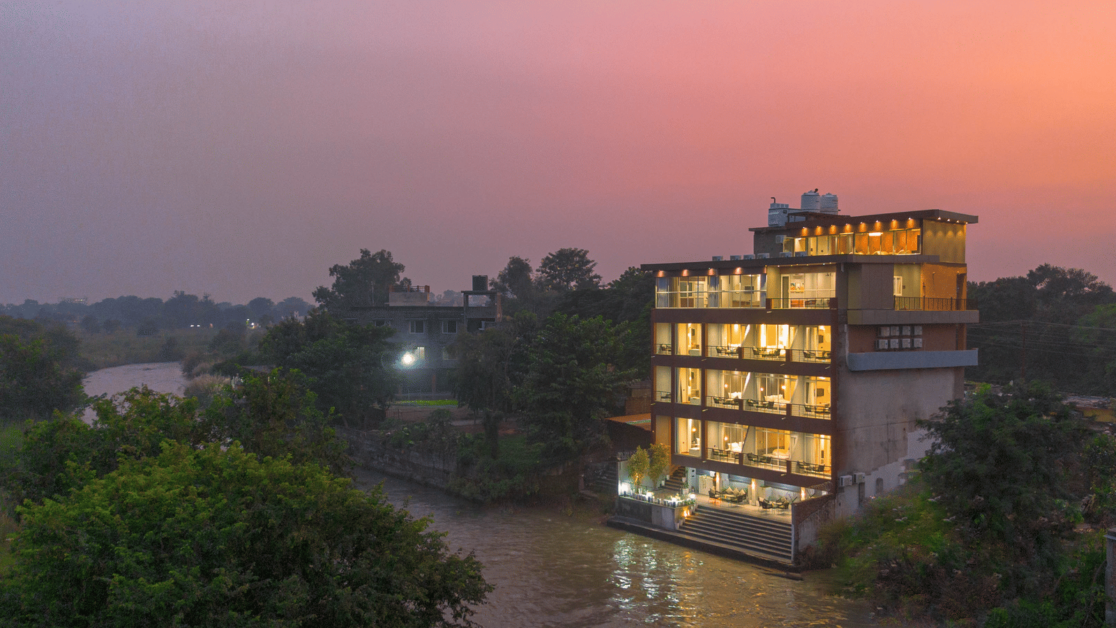 The illuminated Sukoon by the Ganges hotel is captured at sunset, sitting gracefully by the brown-toned river water.