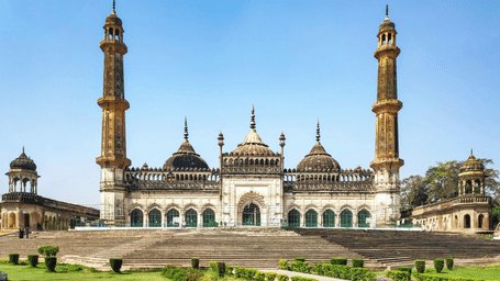 An overview of Bara Imambara in Lucknow with steps leading to the building, two other buildings next to it, a lawn area in the foreground and blue sky in the background.