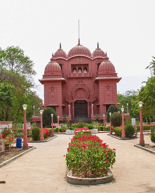 The symmetrical temple at Ramakrishna Ashrama, Rajkot, featuring multiple domes and a carved entrance, with a path lined with flowerbeds and lamp posts.