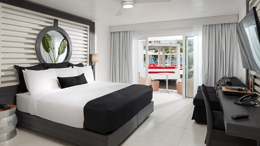 A hotel room in Montego Bay with a plush bed, a wall-mounted television and an outdoor sitting area seen through the window - S Hotel Montego Bay, Jamaica