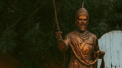 A golden statue of the warrior Shivaji, holding a sword and wearing a turban. The statue stands outdoors at sunset.