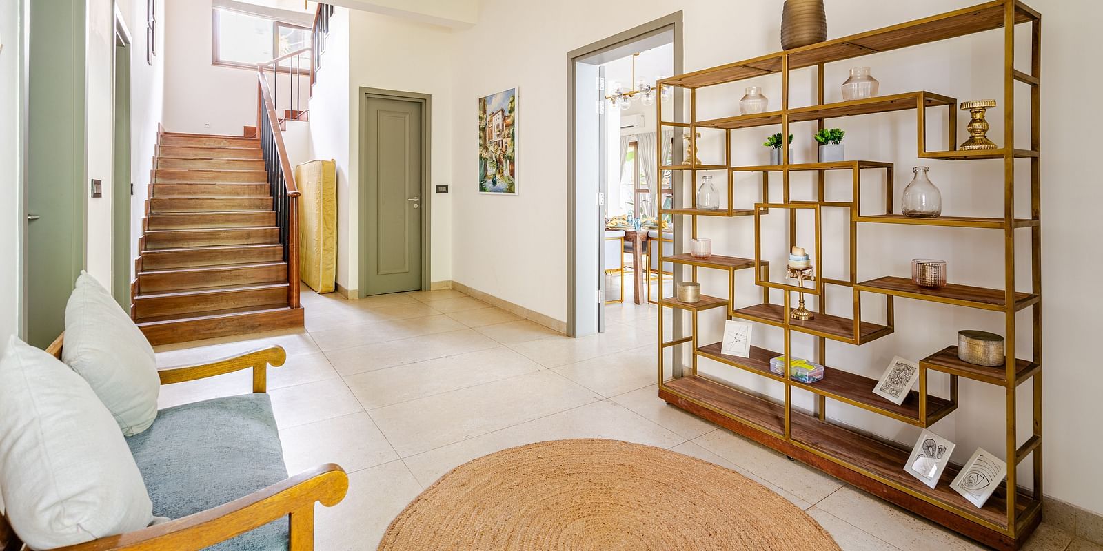 Spacious hallway with a wooden staircase, decorative open-shelf unit, cushioned bench, and natural light streaming in from a large window above - Lindsay Manor