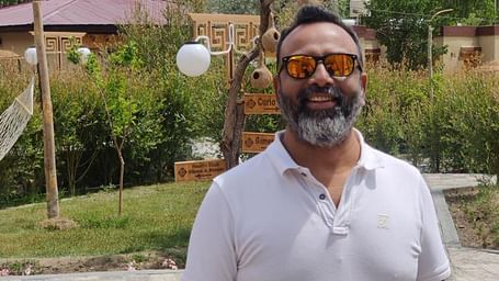 A smiling man with a full beard and orange mirrored sunglasses stands outdoors in a white polo shirt and jeans, with a garden, a building, tall trees, and mountains in the background.