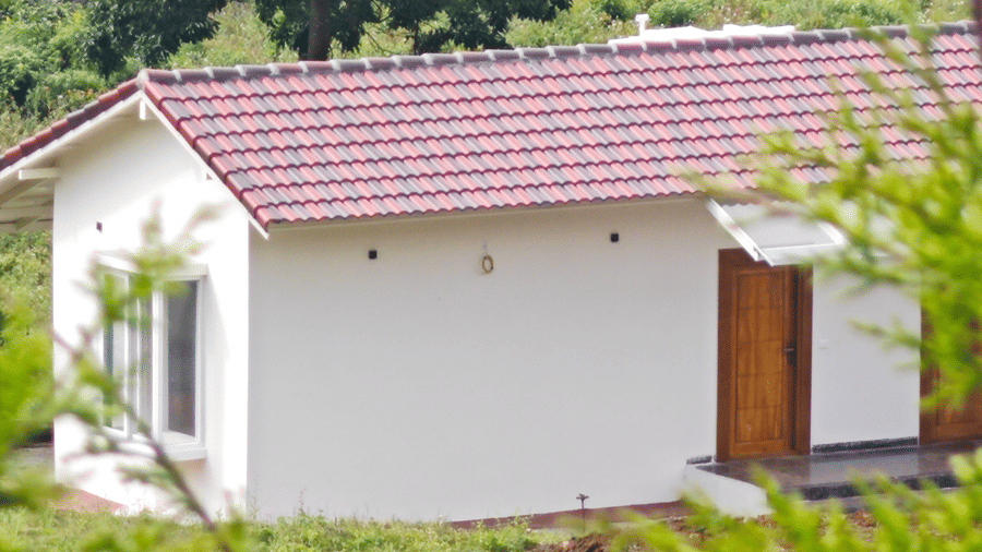 An exterior view shows a single-storey building with a tiled roof and a doorway entrance. Trees and vegetation surround the structure. This building forms part of the Panther accommodation at Ibex Resort, Kotagiri.