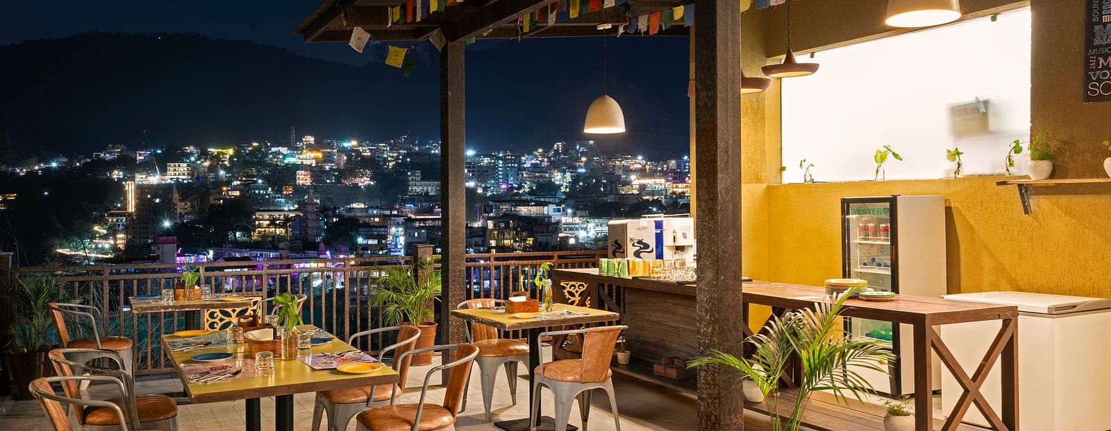 Panoramic view of city skyline from rooftop restaurant, with tables, chairs, and city lights at night at Bedzzz, Rishikesh 