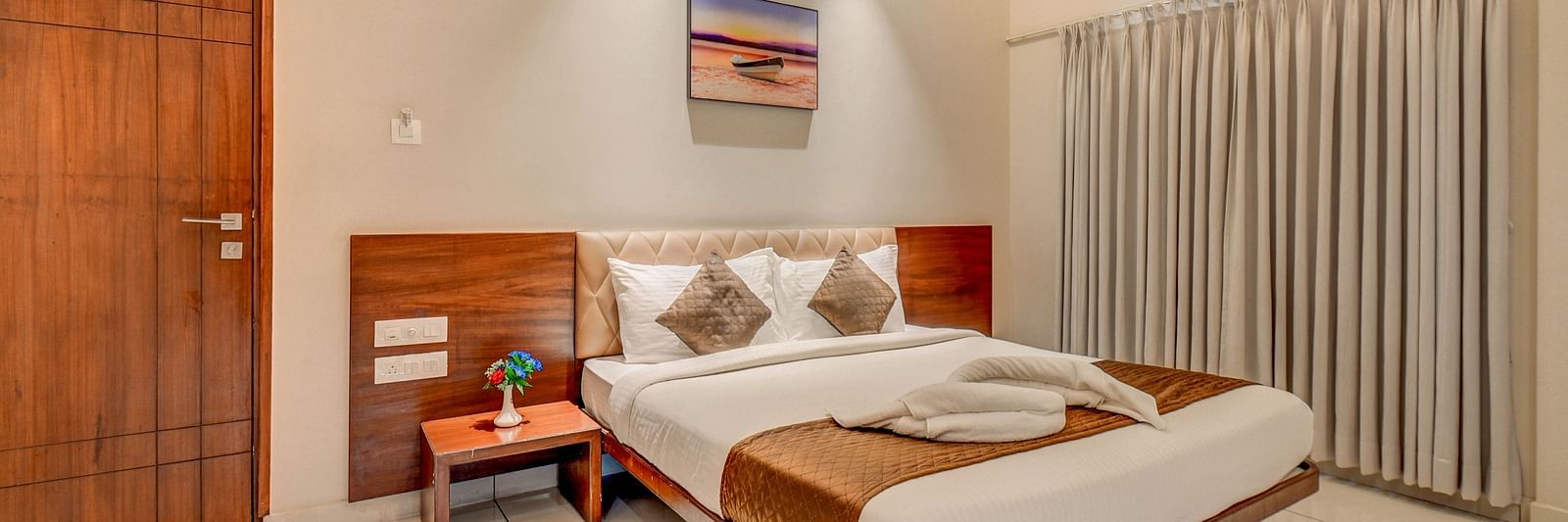 A spacious room at Harigovinda's JNStay with a large bed, wooden headboard, bedside table with a plant, a door, and curtains covering the window