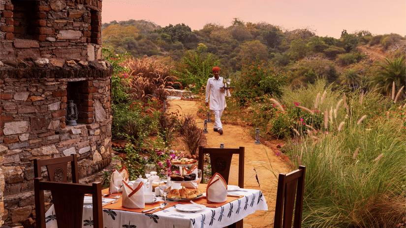 A table for 4 set up next to a stone structure surrounded with greenery and a beautiful sky with pink hues in the background - Chunda Shikar Oudi, Udaipur.